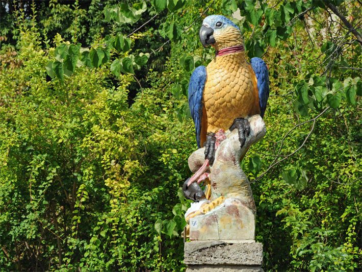 Orange-blau bemalte Papagei-Gartenfigur aus Porzellan im Botanischen Garten München, Veröffentlichung mit freundlicher Genehmigung des Botanischen Gartens München-Nymphenburg
