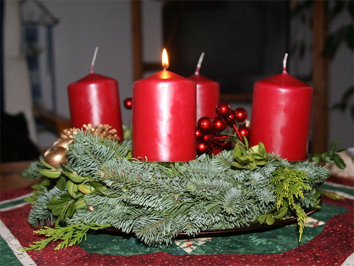 Adventskranz mit Tannenzweigen und vier roten Kerzen am 1. Advent mit einer brennenden Kerze