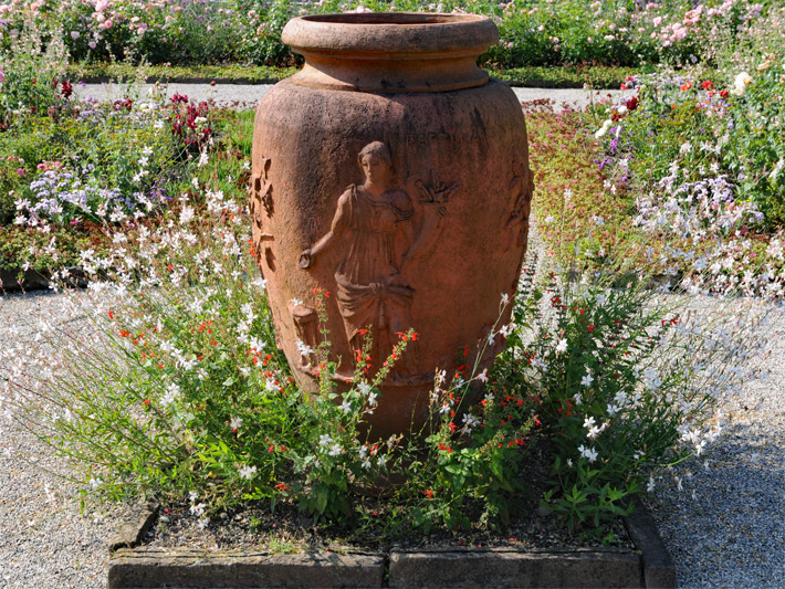 Antike Terracotta-Gartenvase verziert mit einem Motiv aus der griechichen Mythologie einer Göttin in einem Ziergarten mit Sommerblumen