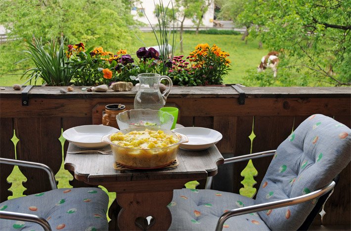 Orange, rote und lila Blumen und grüne Gräser in einem Blumenkasten auf einem Holz-Balkon eines bayerischen Landhauses mit zwei Balkonstühlen und Polster sowie einem gedeckten Balkontisch mit Wasser-Glaskrug, hell-brauner Keramiktasse, weißen Porzellan-Tellern, Keramik-Kochtopf mit Kartoffel-Käse-Gratin und Glasschüssel mit Gurkensalat