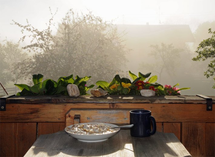 Balkontisch mit einem weißen Teller mit Müsli und einer schwarzen Tasse, dahinter ein bepflanzter Balkonkasten auf einem bayerischen Balkon mit nebeliger Morgenstimmung bei Sonnenaufgang