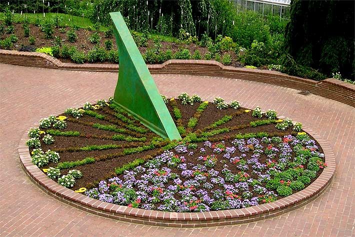 Rundes Blumenbeet mit einer Sonnenuhr-Skulptur, bei dem die Blumen als Ziffernblatt angepflanzt sind