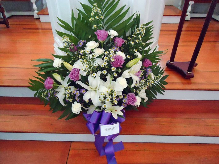 Blumengesteck mit violetten Rosen, weißen Lilien, Margeriten und Gräsern auf einer Holzstufe vor einem Altar in einer Kirche