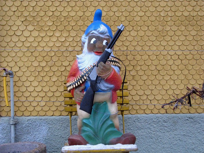Moderner Anti-Gartenzwerg mit Gewehr und Patronengürtel, weißem Bart und bekleidet mit blauer Mütze, ohne Hose und roter Jacke
