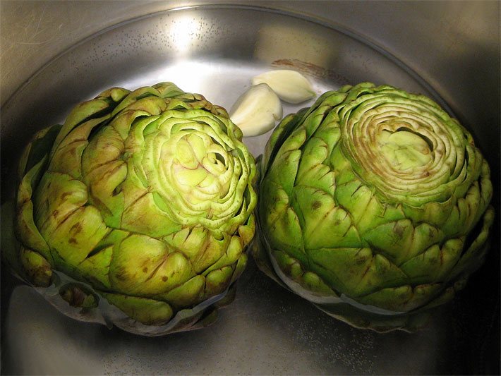 Foto von zwei Artischocken, die in einem silbernen Kochtopf zusammen mit zwei geschälten Knoblauch-Zehen gekocht werden