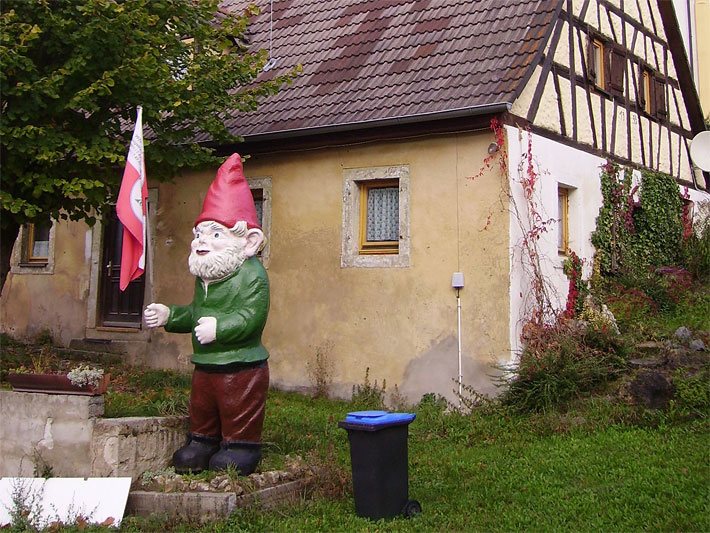Großer Zwerg mit roter Mütze, grünem Hemd und brauner Hose als XXL-Standfigur mit Fahne vor einem Fachwerkhaus