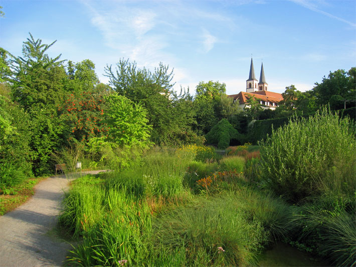 Das Gelände der Landesgartenschau Würzburg 1990 mit Bäumen, Sträuchern und einem Weg mit Rastbank