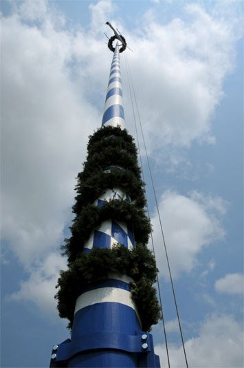 Maibaum in Bayern mit blau-weissem Rautenmuster sowie Girlanden am unteren Ende und einem Kranz am oberen Ende aus Nadelzweigen