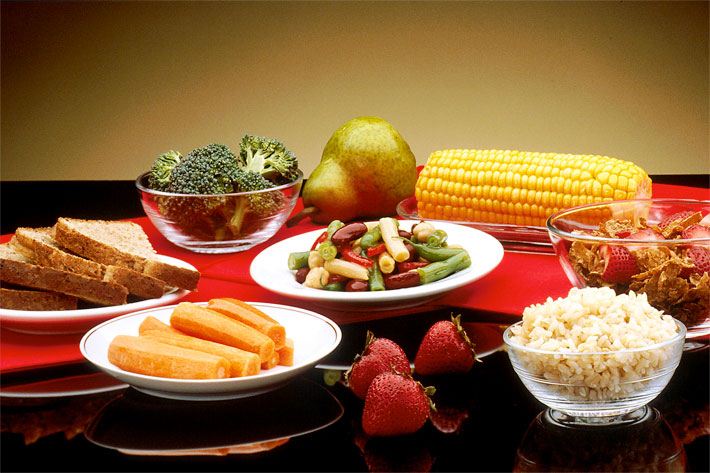 Verschiedene Teller mit geschnittenem Brot und verschiedenen Getreide-, Obst- und Gemüsesorten wie Brokkoli, Bohnen, Reis, Karotten/Rüben sowie einer Birne, Erdbeeren und einem Maiskolben