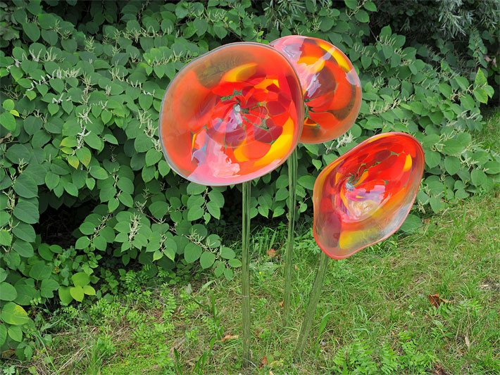 Drei orange-rote trichterförmige Glasblumen mit gläsernen, über 50 cm hohen Blumenstiehlen als Gartendeko