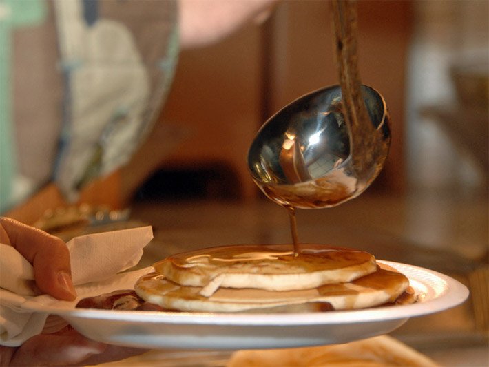 Pancakes, auch Pfannkuchen oder Eierkuchen, auf einem weissen Teller, die mit einer Schöpfkelle mit Ahornsirup übergossen werden