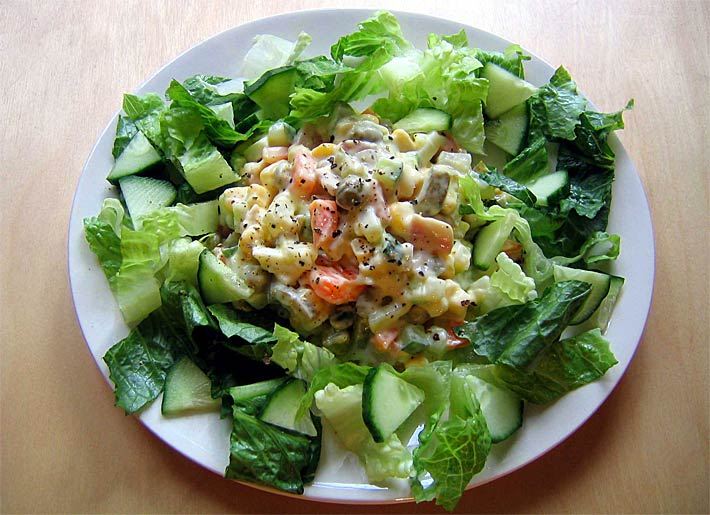 Russischer Salat auf Blattsalat mit Gurken, Myonnaise, Kartoffeln, Eier-, Fisch- und Wurststücken auf einem weißem Teller