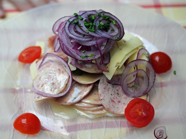 Schweizer Wurstsalat auf einem Glasteller garniert mit Wurst-Scheiben, Käse-Streifen, Zwiebel-Ringen, geschnittenem Schnittlauch und einem Dressing aus Essig und Öl