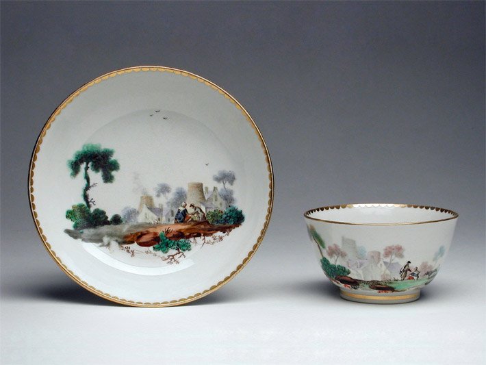 Weiße  Porzellan-Teetasse mit Goldrand und einer aufgestellten weißen Porzellan-Untertasse mit Goldrand, die jeweils bemalt sind mit einer Frau und einem Mann auf dem Land im Gespräch