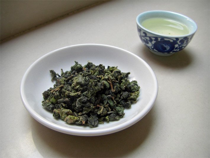 Weiße Porzellan-Schale mit Teeblättern und einer typisch chinesischen, weißen Porzellan-Trinktasse ohne Henkel mit blauer Blumen-Bemalung, wobei sich beide Geschirr-Stücke auf den Tieguanyin-Tee beziehen, einer Oolong-Sorte